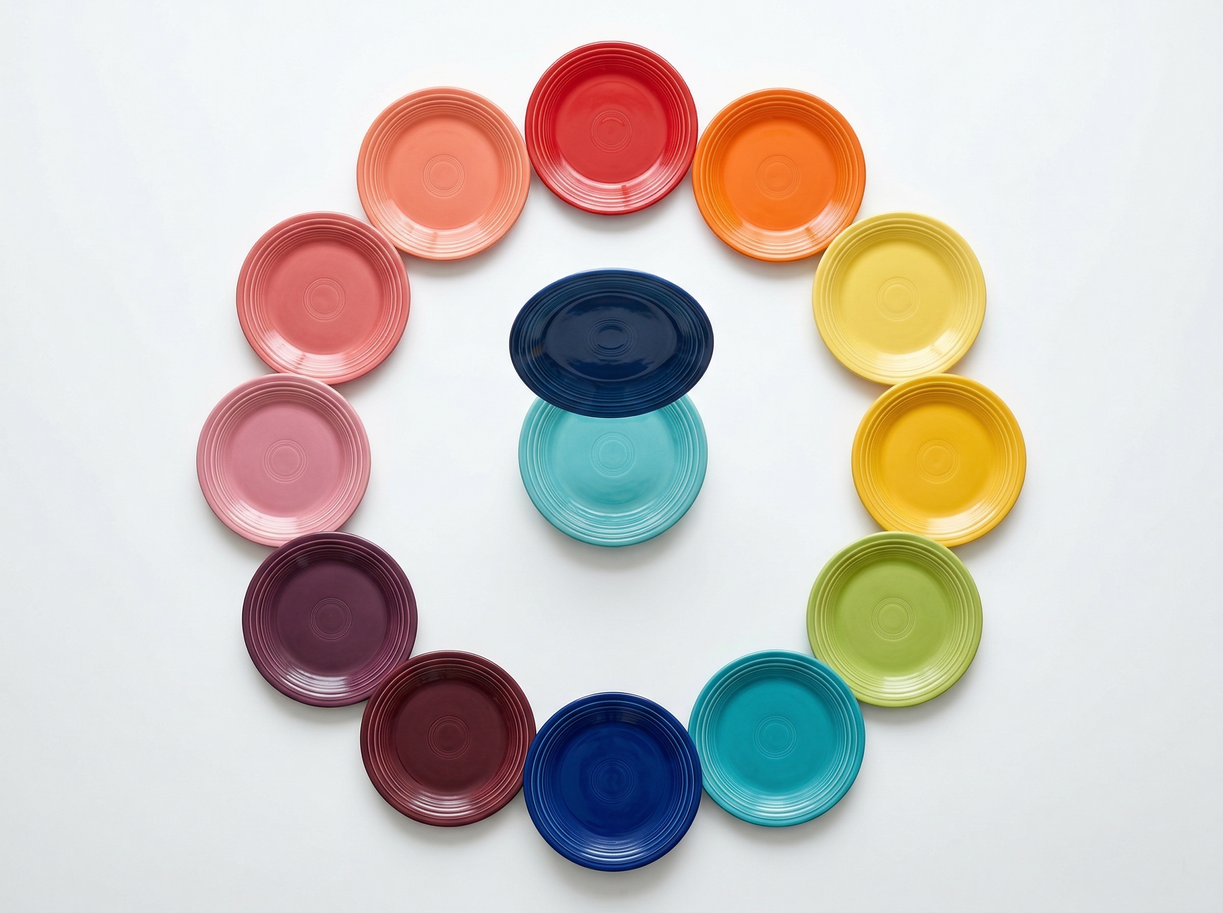 Fifteen Fiesta plates arranged in a color wheel circle viewed from above, with one plate being removed